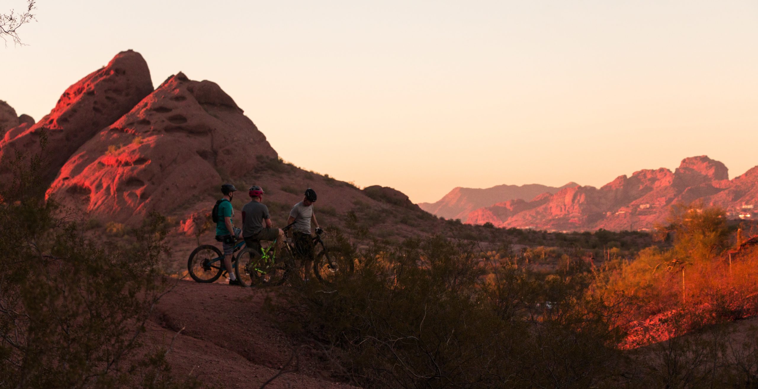 3 people riding bikes at sunset with mountains in the background and red rock all around while on a mountain bike tour.