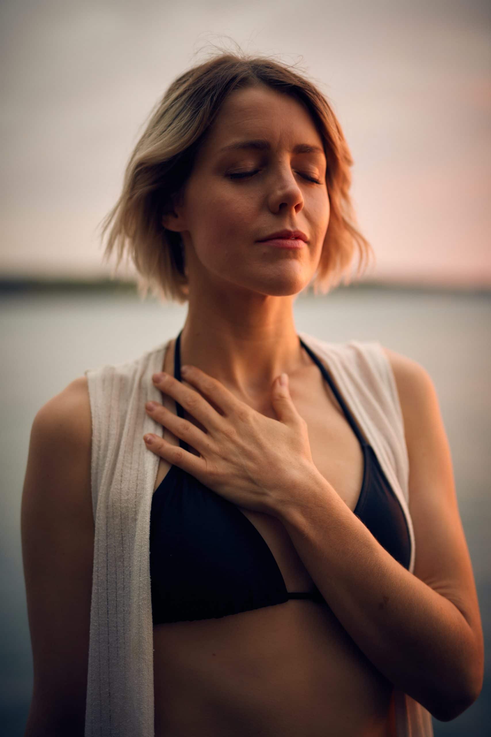 Woman practicing breathing exercises and mindfulness with hand over her heart in black swimsuit and white jacket