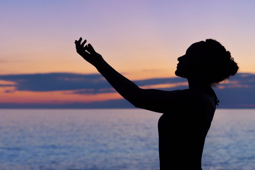 Indigenous Black Woman practicing mindfulness and raising arms over the ocean at sunset 