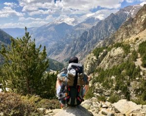 Man with a backpack practicing mindfulness while looking at a valley, trees, and mountains in the distance.