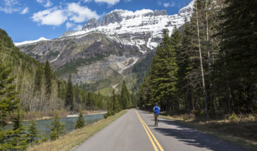A biker on an empty roading going through Glacier National Park with a lake on the left and snow covered mountains ahead