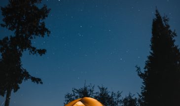 A lit tent camping under a dark sky full of stars with water and trees behind it in the Boundary Waters Canoe Area