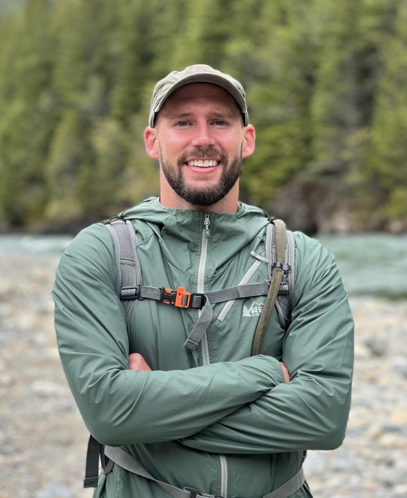 Man in green jacket standing in front of a river, trees and mountains in the background.