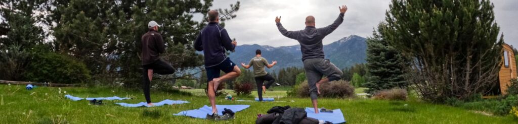 People doing yoga in front of mountains in the background with green grass and green trees.