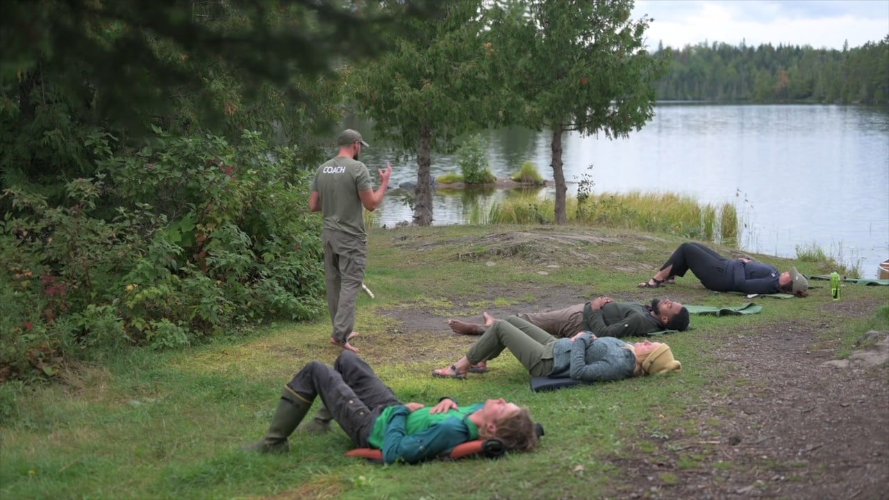 A man coaching breathwork with participants lying on the ground in a forest near a lake