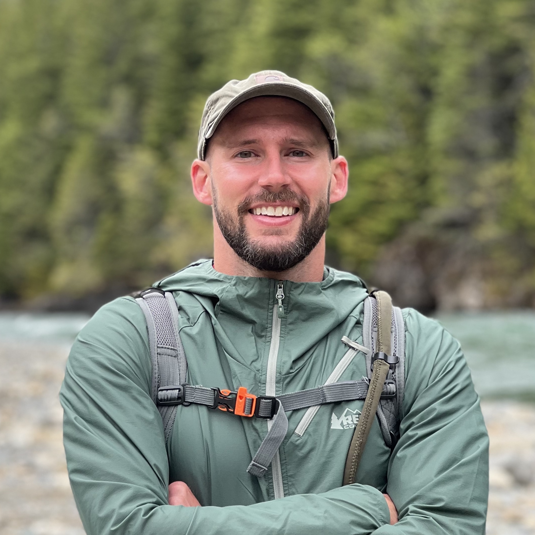 Man in a green jacket standing with his arms folded and a backpack on in front of a river and green pine trees.