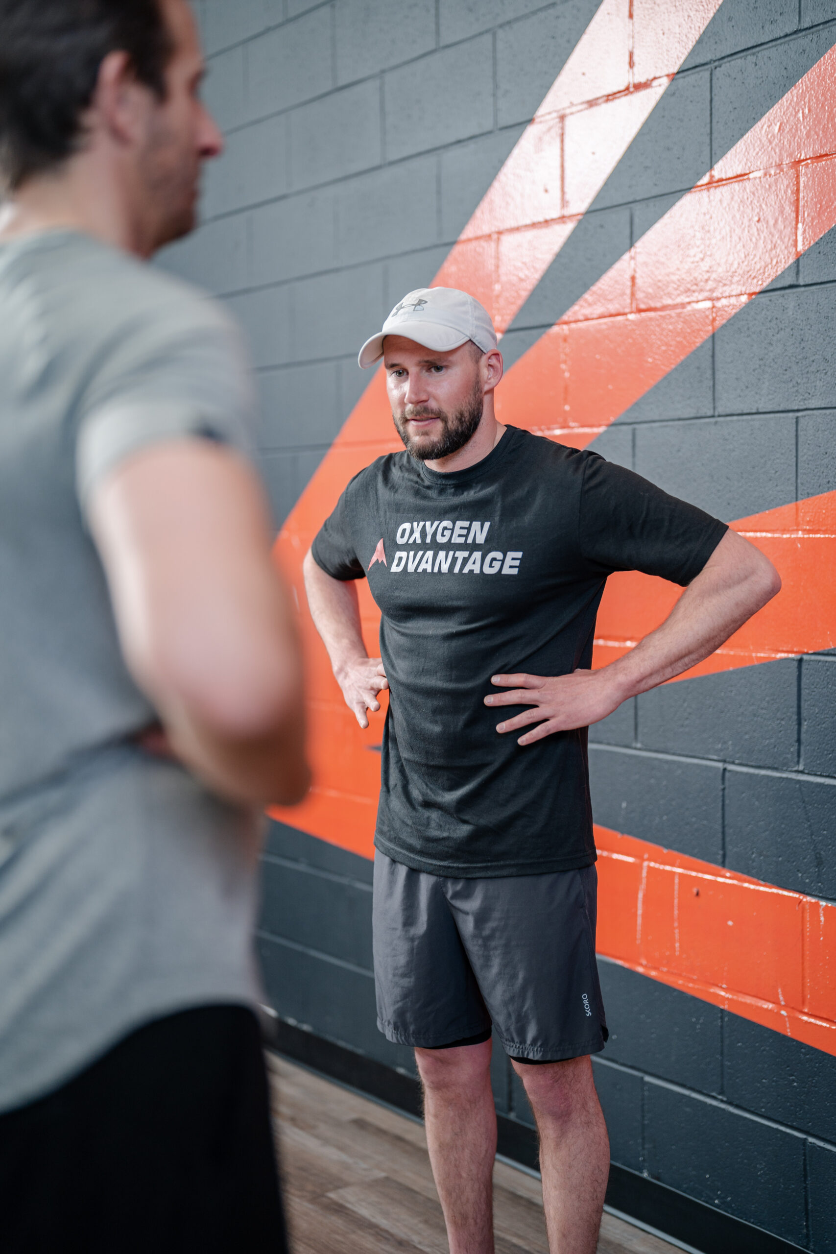 Man wearing a black shirt that says Oxygen Advantage on it has his hands on ribs and coaching someone on a breathwork exercise at a gym.