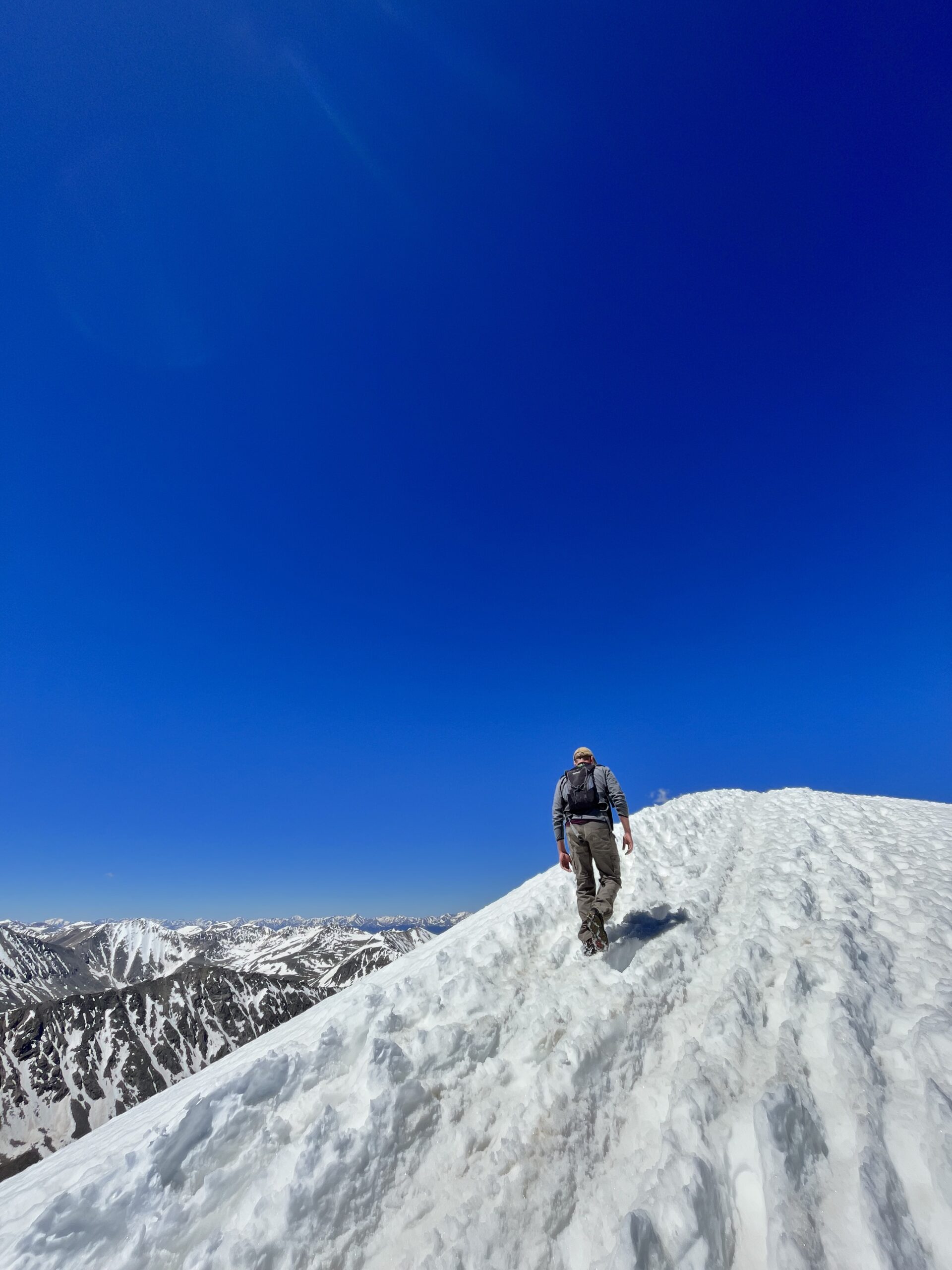 Man walking on a snowy trail to a mountain peak with a blue sky and mountains in the distance.